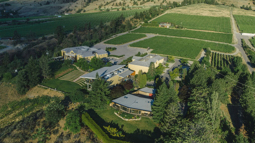 Aerial view of a vineyard estate with multiple buildings surrounded by lush green fields and trees. The landscape includes well-maintained gardens, pathways, and a backdrop of rolling hills under a clear sky.